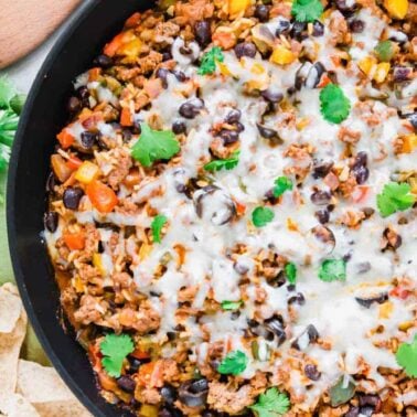 A skillet filled with cooked ground beef, black beans, diced vegetables, and melted cheese, garnished with cilantro—this easy dinner is perfect for anyone seeking satisfying meals. Served alongside tortilla chips and fresh cilantro.