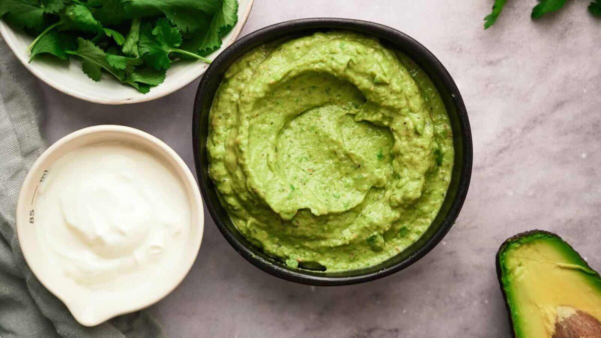 A bowl of green avocado dip, a flavorful gluten-free sauce, sits next to sour cream, fresh cilantro, and half an avocado on a light surface.
