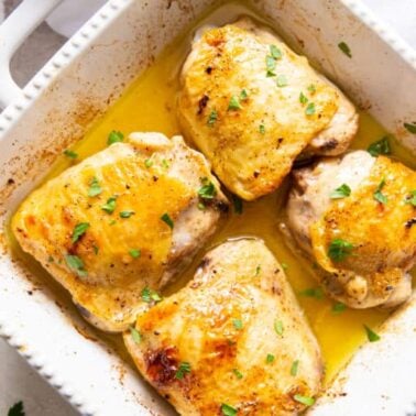 Four baked chicken thighs with golden skin in a white ceramic baking dish, garnished with chopped parsley and sitting in a pool of juicesโperfect for low carb dinners that are both satisfying and delicious.