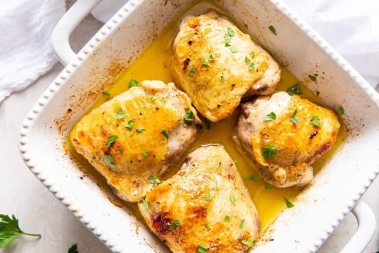 Four baked chicken thighs with golden skin in a white ceramic baking dish, garnished with chopped parsley and sitting in a pool of juices—perfect for low carb dinners that are both satisfying and delicious.