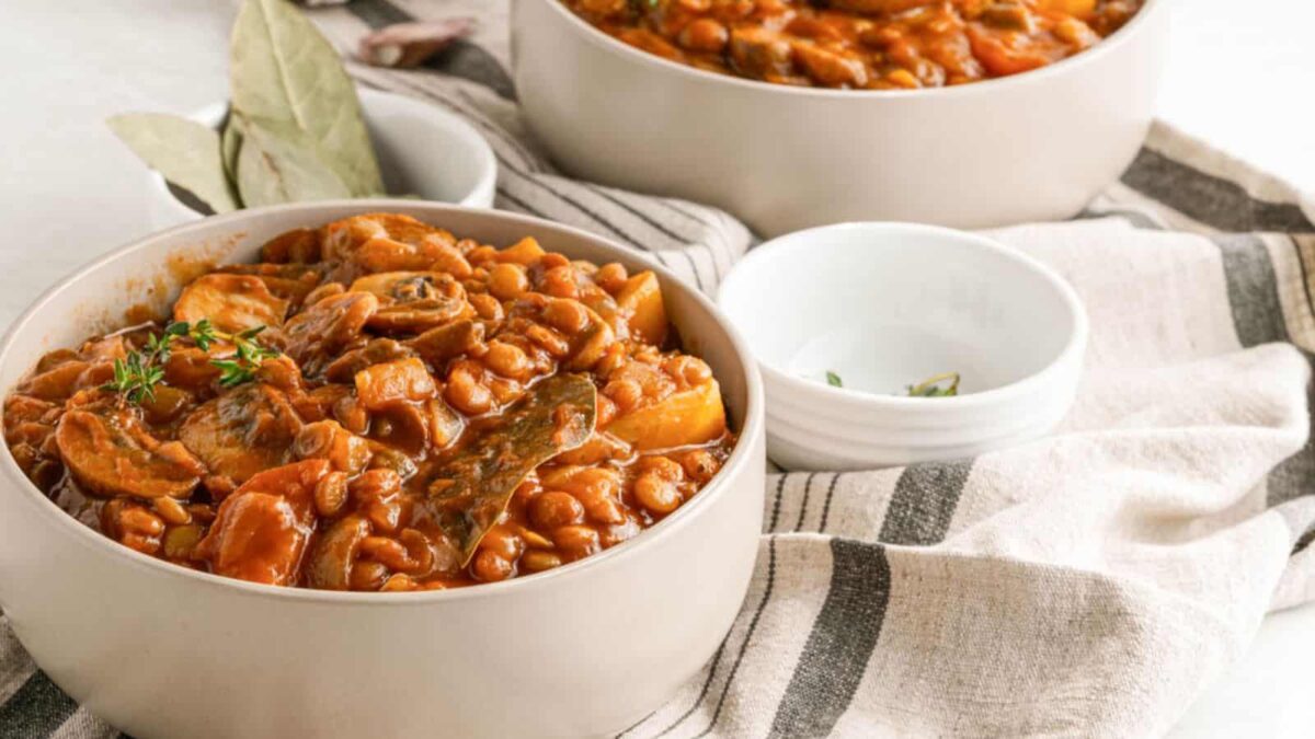 Two bowls of baked beans with mushrooms and herbs sit on a striped cloth, next to empty small white bowls—a hearty comfort food perfect for fans of classic homestyle stew recipes.