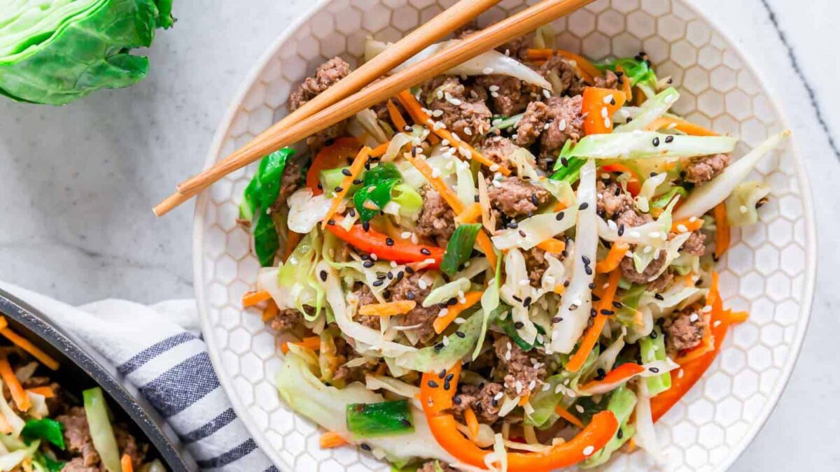 A bowl of stir-fried ground meat, shredded cabbage, sliced red bell peppers, carrots, and green onions, topped with black and white sesame seeds—classic comfort food that's perfect for mains and sides. Chopsticks rest on the bowl.