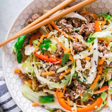 A bowl of stir-fried ground meat, shredded cabbage, sliced red bell peppers, carrots, and green onions, topped with black and white sesame seedsโclassic comfort food that's perfect for mains and sides. Chopsticks rest on the bowl.