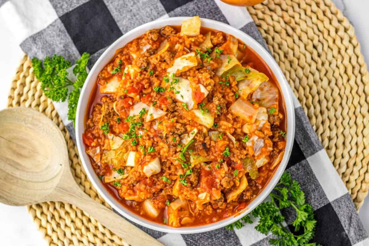 A bowl of unstuffed cabbage roll casserole with ground meat, cabbage, and tomato sauce, garnished with parsley—a perfect comfort food option among cabbage dishes, served on a woven placemat with a wooden spoon nearby.