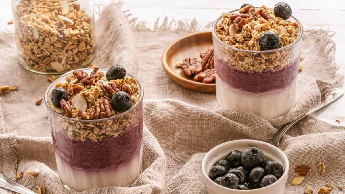 Two glasses of layered parfait with yogurt, blueberry puree, crunchy clusters from granola recipes, and pecans sit on a cloth, with a small bowl of blueberries and a plate of pecans nearby—perfect for breakfast.