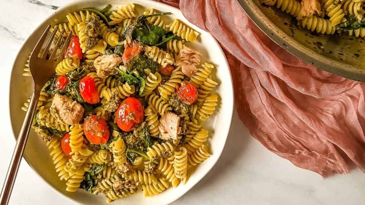 A bowl of rotini pasta with cherry tomatoes, spinach, mushrooms, basil, and chunks of tuna, served with a fork on the side.