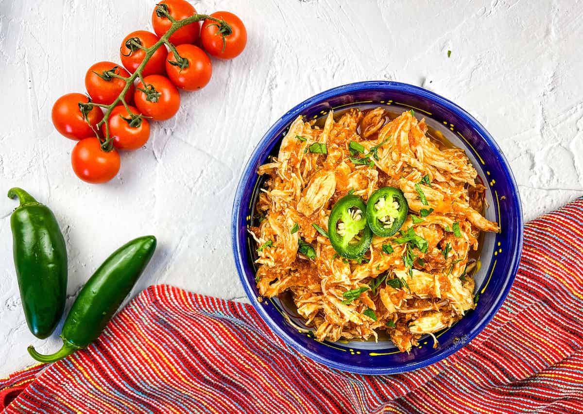 A bowl of juicy meat shredded chicken topped with sliced jalapeños and herbs, next to cherry tomatoes, jalapeños, and a striped red cloth on a white surface. Perfect for satisfying Instant Pot chicken dinners.