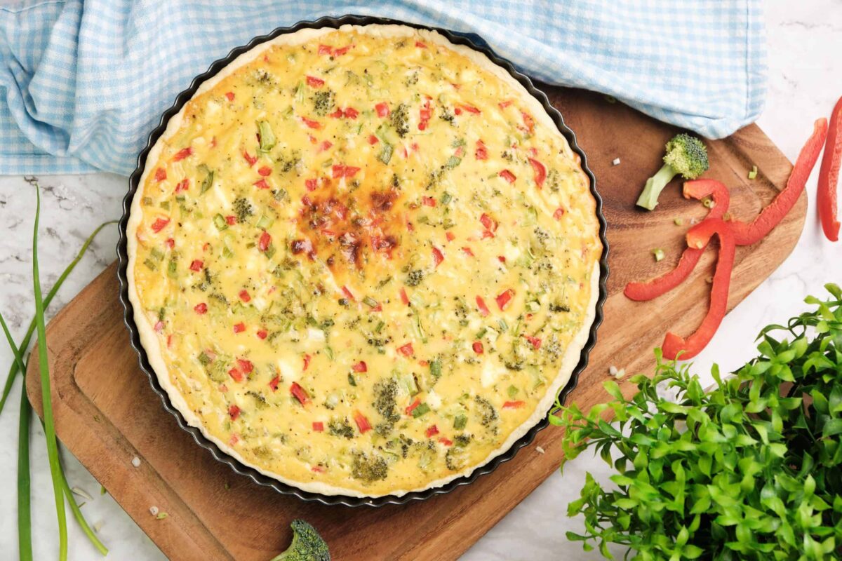 A baked vegetable quiche with savory fillings of broccoli and red bell peppers sits on a wooden board, next to a blue checkered cloth and fresh greens.