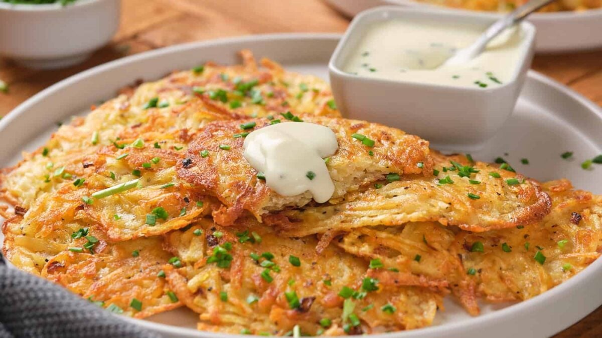 A plate of golden-brown potato pancakes, reminiscent of flavorful bites from classic fritter recipes, garnished with chopped chives, topped with a dollop of sour cream, and served with a small bowl of dipping sauce.