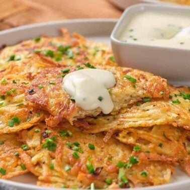A plate of golden-brown potato pancakes, reminiscent of flavorful bites from classic fritter recipes, garnished with chopped chives, topped with a dollop of sour cream, and served with a small bowl of dipping sauce.