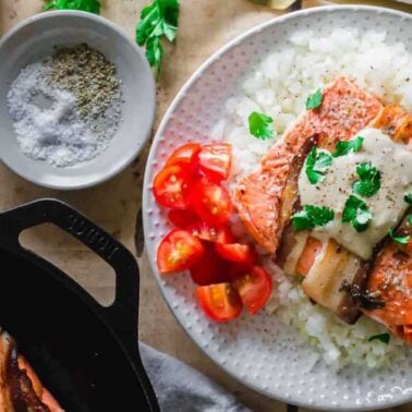 A plate of cooked salmon fillets with melted cheese, crispy bacon, and herbs, served over rice with sliced cherry tomatoes. Salt, pepper, and herbs are on the side.