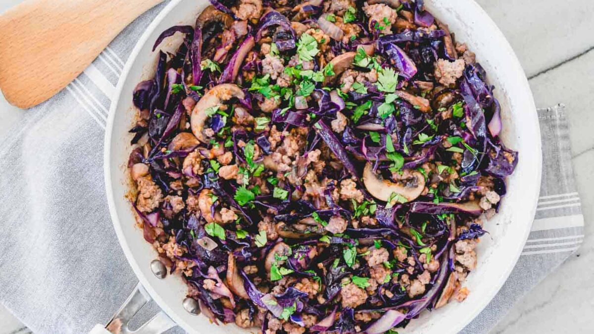 A skillet filled with cooked ground meat, sliced mushrooms, and shredded red cabbage, offering rich savory notes and garnished with chopped parsley—perfect for fans of hearty cabbage dishes.
