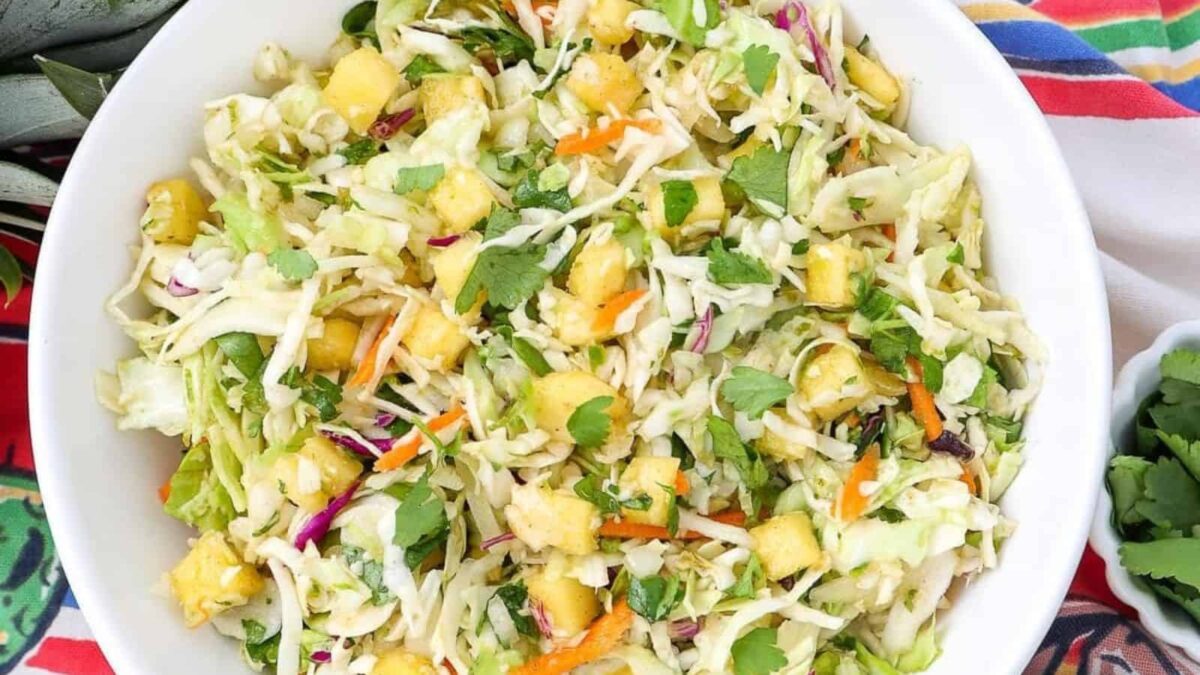 A bowl of cabbage slaw features tender leaves, pineapple chunks, shredded carrots, cilantro, and other vegetables—perfect for those who love vibrant cabbage dishes with subtle savory notes.