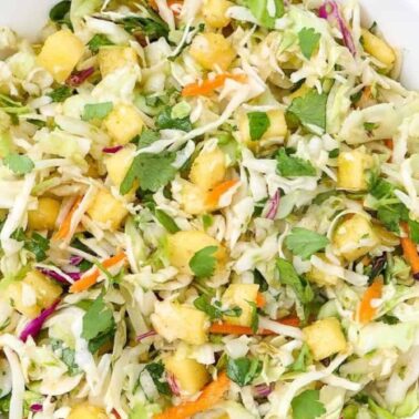 A bowl of cabbage slaw features tender leaves, pineapple chunks, shredded carrots, cilantro, and other vegetablesโperfect for those who love vibrant cabbage dishes with subtle savory notes.