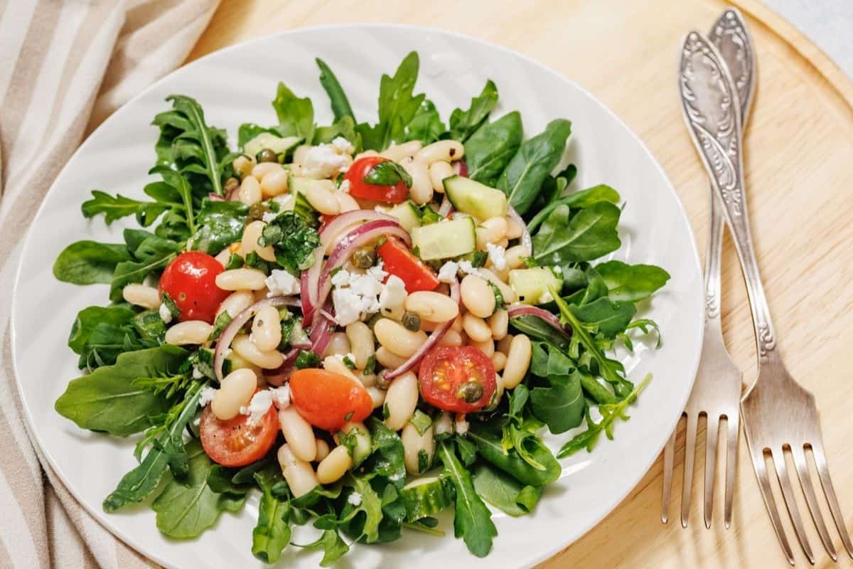 A plate of arugula salad with cherry tomatoes, white beans, cucumber, red onion, feta cheese, and herbs—perfect for fresh sides or light mains. Two forks and spoons are placed beside the plate on a wooden surface.