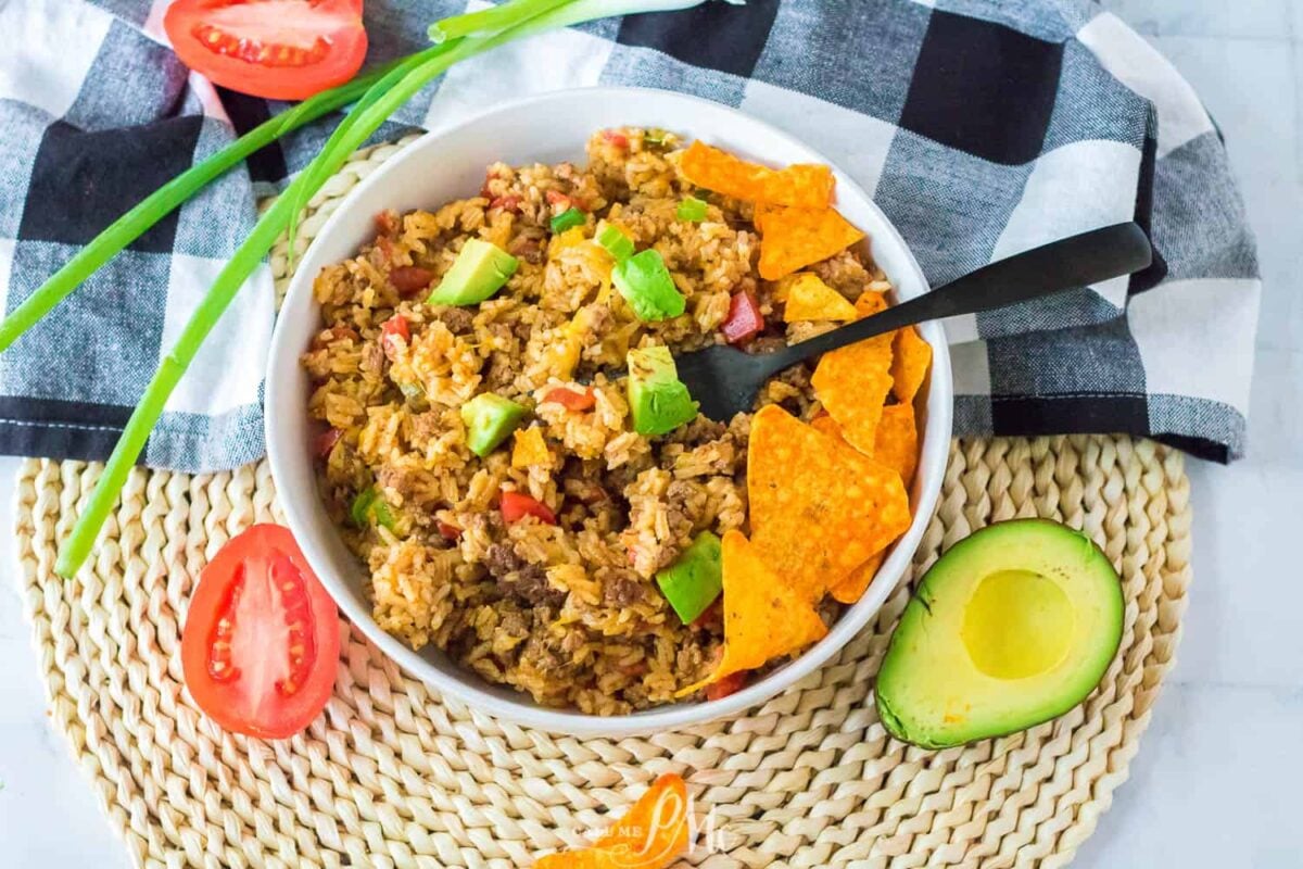 A bowl of Mexican-style rice topped with avocado chunks, tortilla chips, seasoned strips, and sizzling peppers, with tomato slices, green onions, and half an avocado on the side. A black fork rests in the bowl.