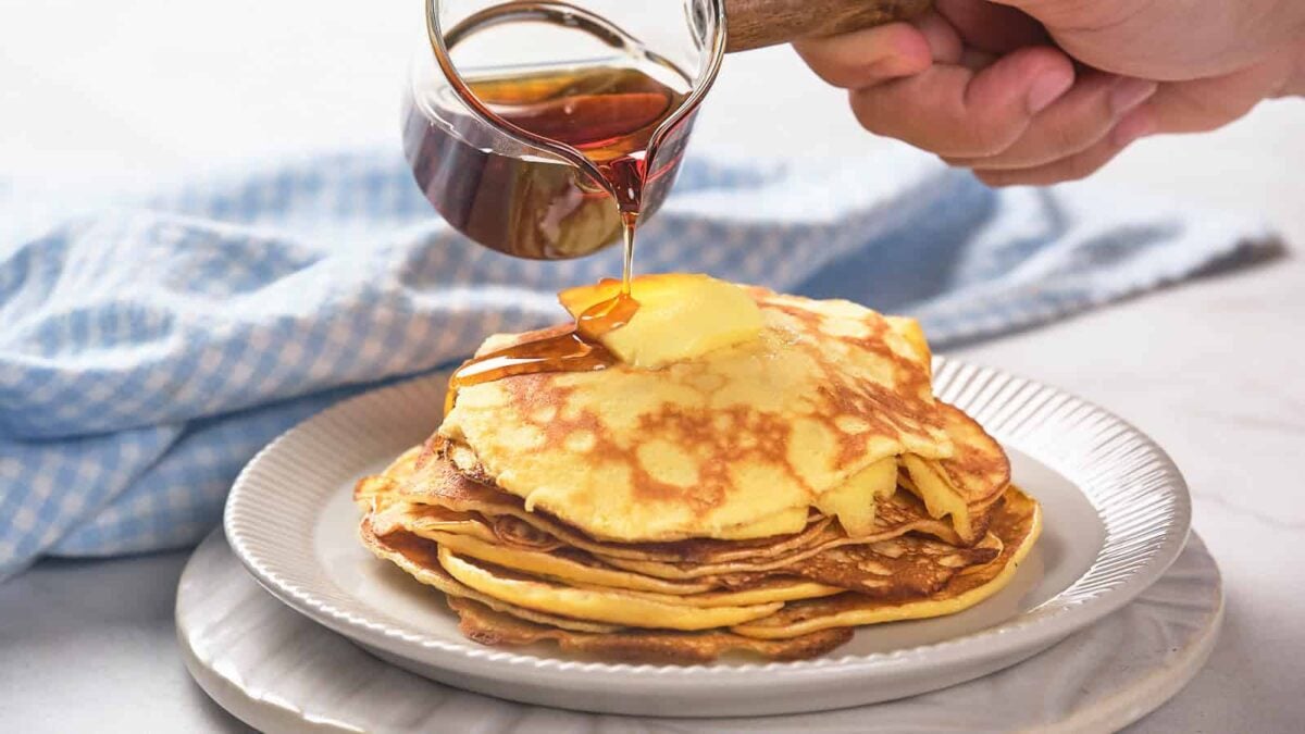 A hand pours syrup onto a stack of fluffy pancakes with golden edges, topped with butter and served on a white plate, set against a blue checked cloth.