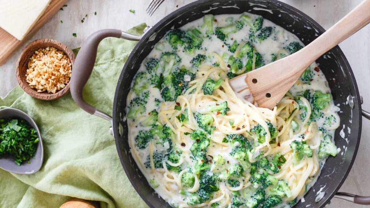 A pan of creamy pasta with broccoli, reminiscent of classic Alfredo recipes, is being stirred with a wooden spoon on a green cloth beside bowls of chopped herbs and toasted breadcrumbs.