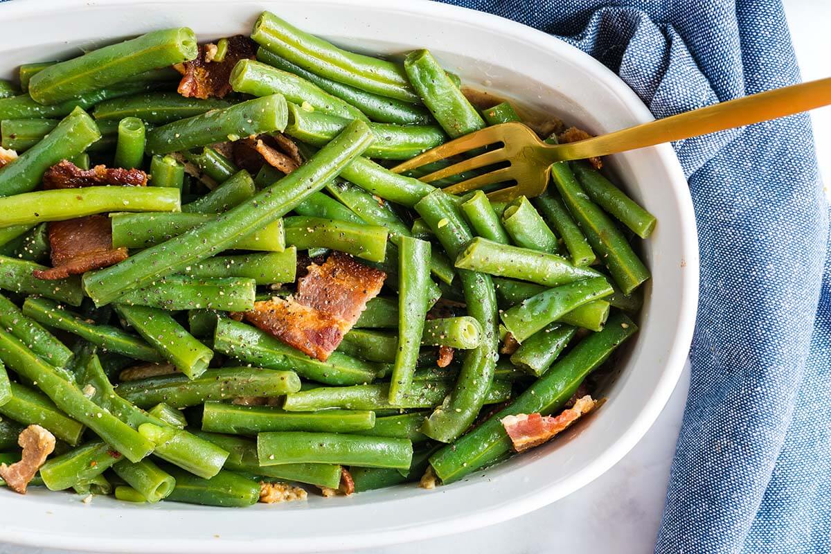Oval dish filled with cooked green beans and pieces of bacon, with a gold fork resting inside; a delicious option for easy sides at weeknight meals. Blue cloth napkin on the side.