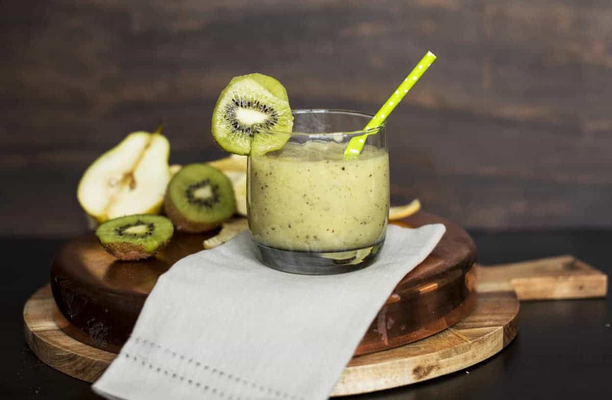 A glass of green smoothie with a straw and kiwi slice garnish sits on a napkin, with sliced kiwi and pear in the background on a wooden board—showcasing the vibrant appeal of fresh smoothies and bright fruits.