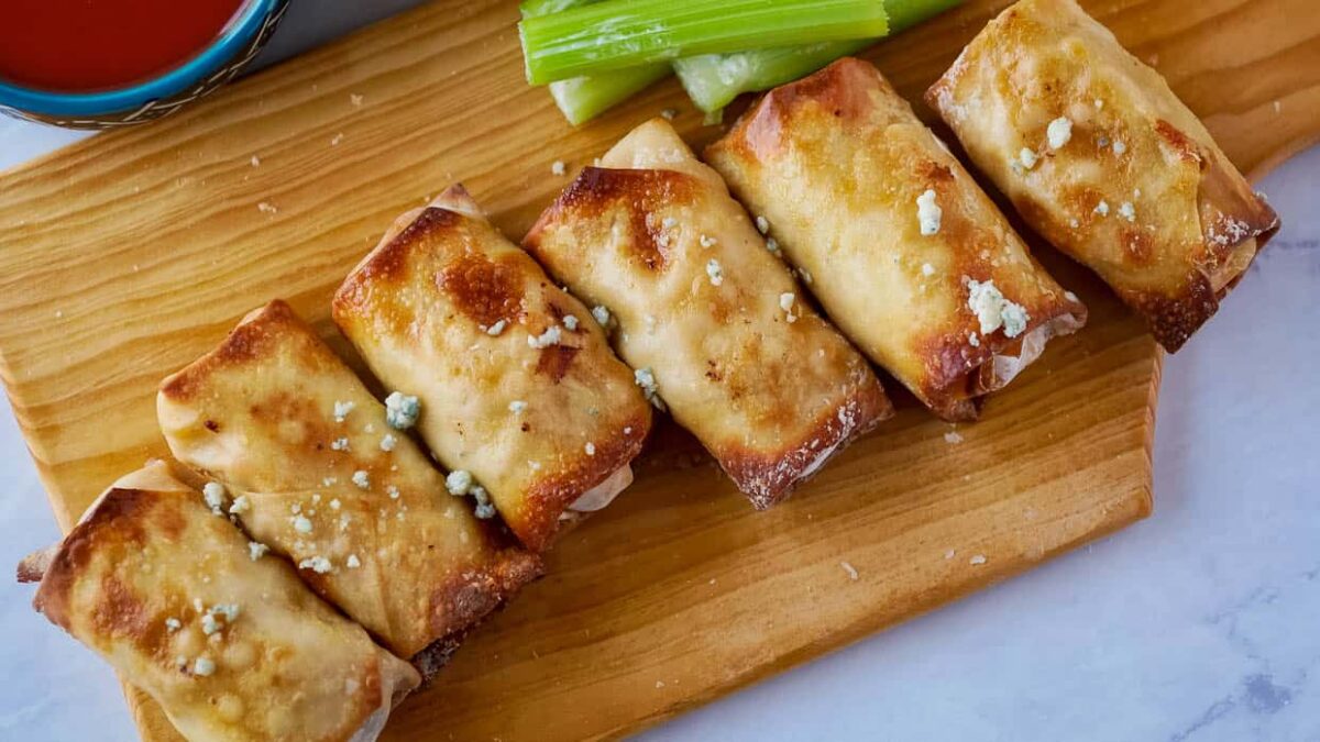 Six golden-brown egg rolls with crunchy fillings are arranged on a wooden board, garnished with crumbled cheese, accompanied by celery sticks and a bowl of flavorful dipping sauces.