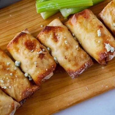 Six golden-brown egg rolls with crunchy fillings are arranged on a wooden board, garnished with crumbled cheese, accompanied by celery sticks and a bowl of flavorful dipping sauces.