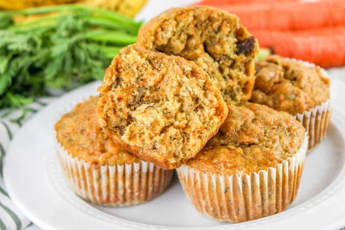 A plate of carrot muffins, known for their high flavor, with one muffin cut in half to show the moist interior, sits on a white plate. Fresh carrots and greens are visible in the background.