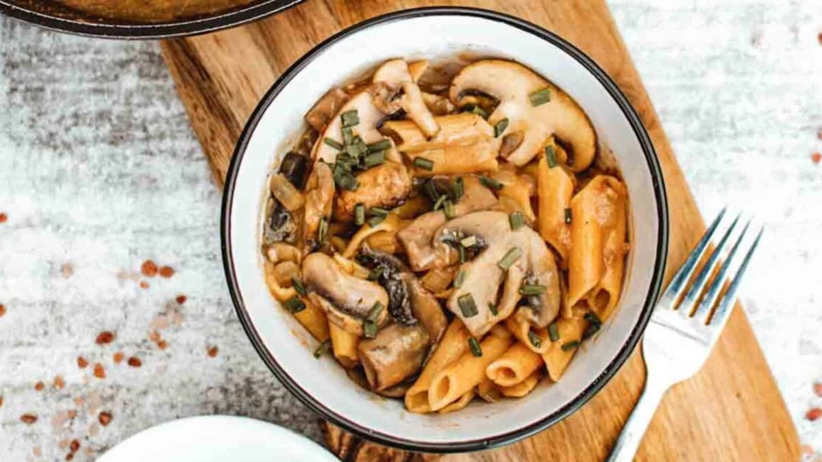 A bowl of penne pasta with sliced mushrooms and herbs on a wooden board, next to a fork—a perfect pick for mushroom dinner recipes brimming with savory flavor.