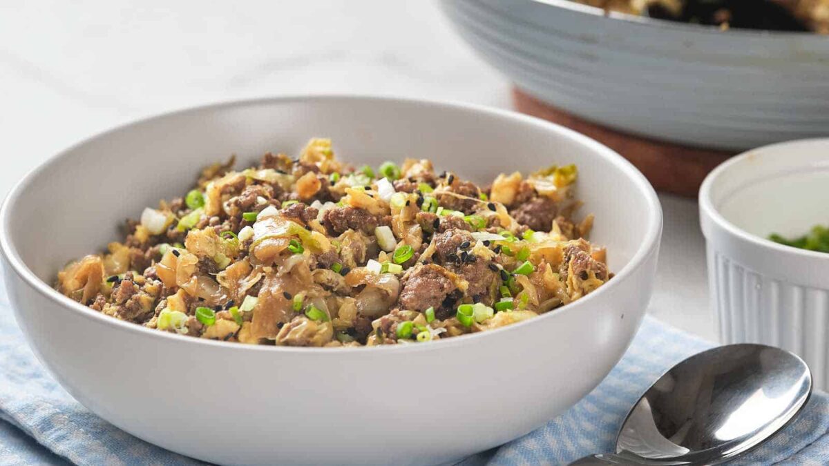 A white bowl filled with a mixture of ground meat, chopped vegetables, green onions, and crunchy fillings sits on a blue cloth next to a spoon—perfect for egg roll ideas or pairing with your favorite dipping sauces.
