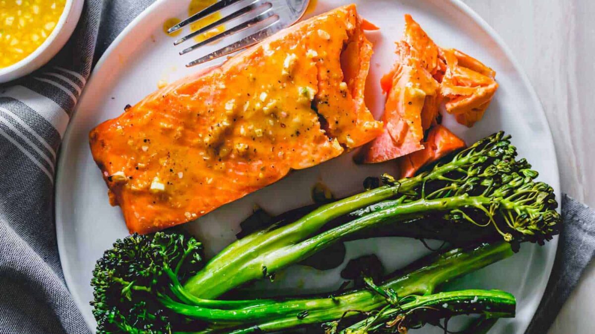 A plate with a piece of cooked salmon topped with a sweet and nutty sauce, steamed broccolini, and a fork—perfect for those who love seasonal recipes.