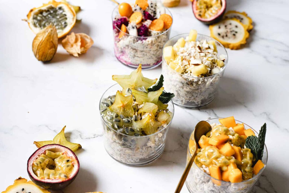 Four glasses of hearty grains oatmeal topped with tropical fruits like starfruit, passion fruit, melon, and berries are arranged on a white marble surface with scattered fruit pieces—perfect for delicious and wholesome bites.