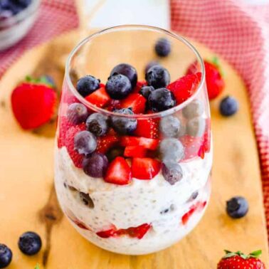 A glass filled with layered overnight oats, hearty grains, strawberries, and blueberries sits on a wooden board, with more berries scattered around and a red checkered cloth in the background.