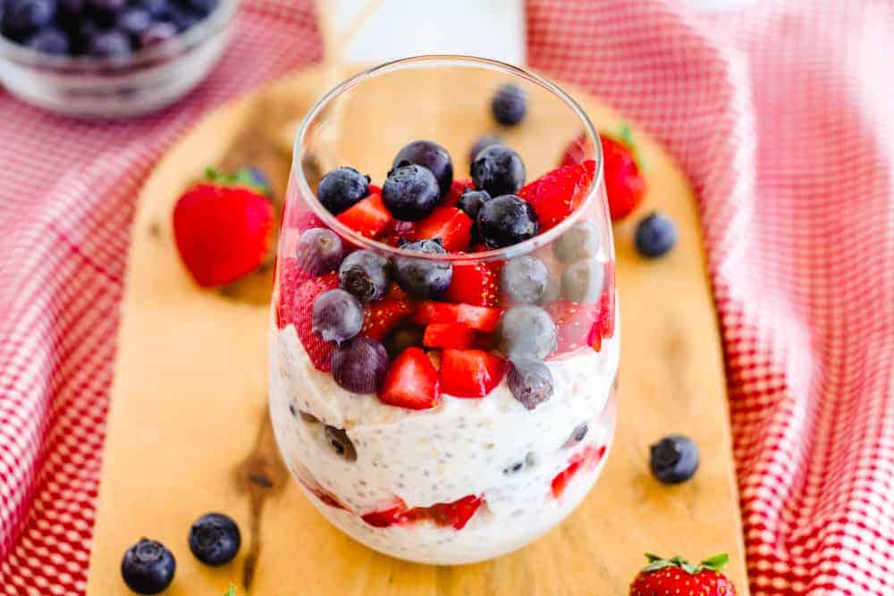 A glass filled with layered overnight oats, hearty grains, strawberries, and blueberries sits on a wooden board, with more berries scattered around and a red checkered cloth in the background.