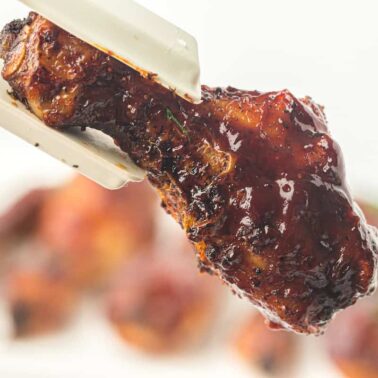 A cooked, juicy chicken drumstick coated in barbecue sauce is held by white tongs against a blurred background, showcasing the irresistible appeal of air fryer chicken.