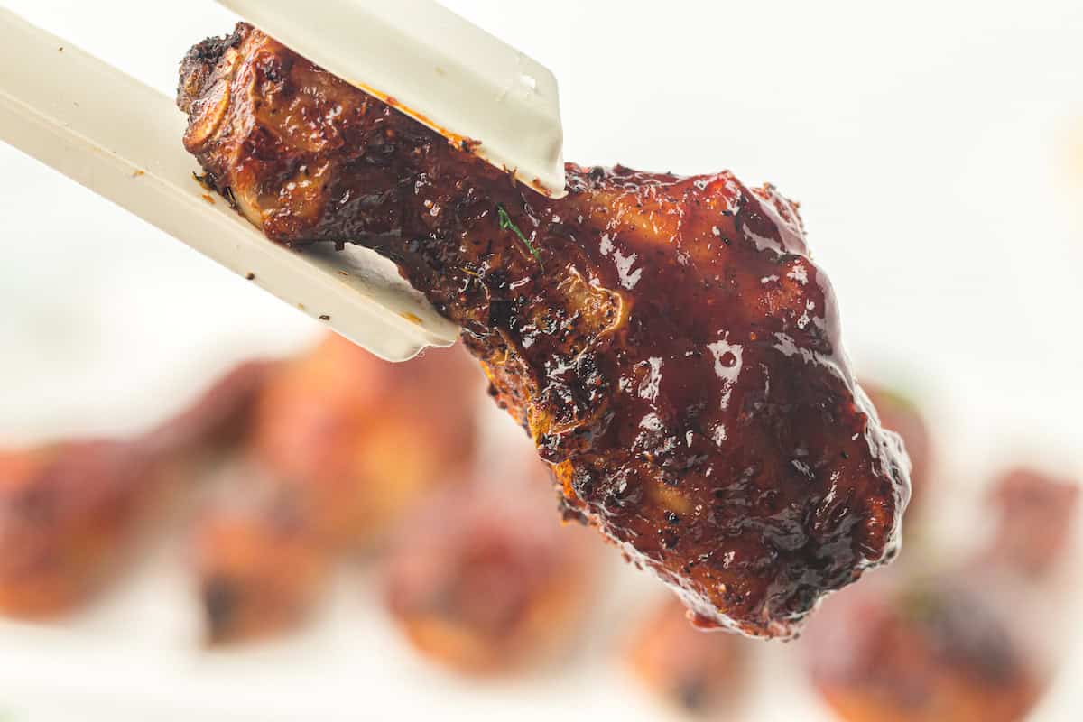 A cooked, juicy chicken drumstick coated in barbecue sauce is held by white tongs against a blurred background, showcasing the irresistible appeal of air fryer chicken.