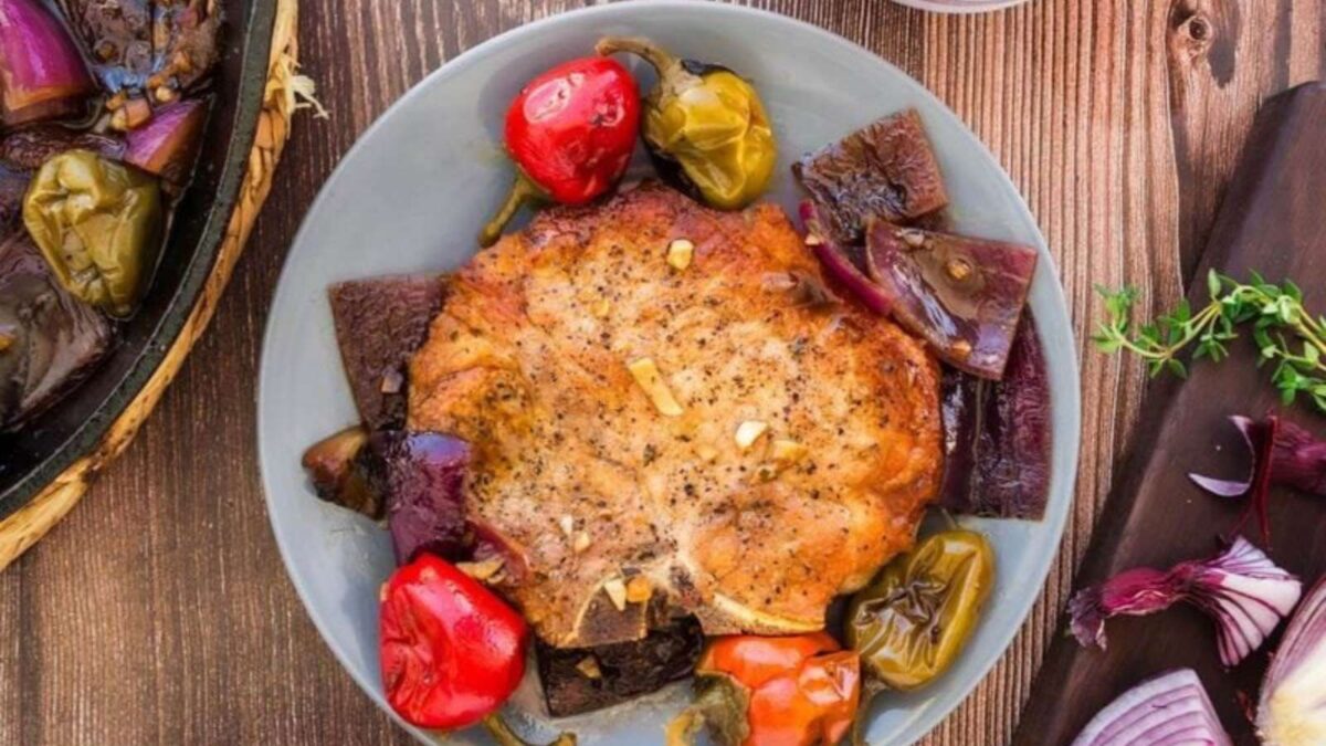 A plate with juicy pork chops surrounded by red and green peppers, red onion pieces, and herbs, seasoned with bold seasoning, sits on a wooden table.