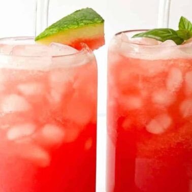 Two tall glasses filled with vibrant sips of iced red watermelon drinks, garnished with a watermelon slice and mint leaves, sit side by side on a white surfaceโrefreshing vegan drinks made with fresh ingredients.