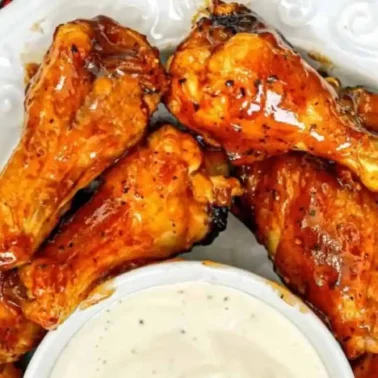 A plate of BBQ chicken wings with a smoky flavor is arranged around a small bowl of creamy dipping sauce on a white dish.