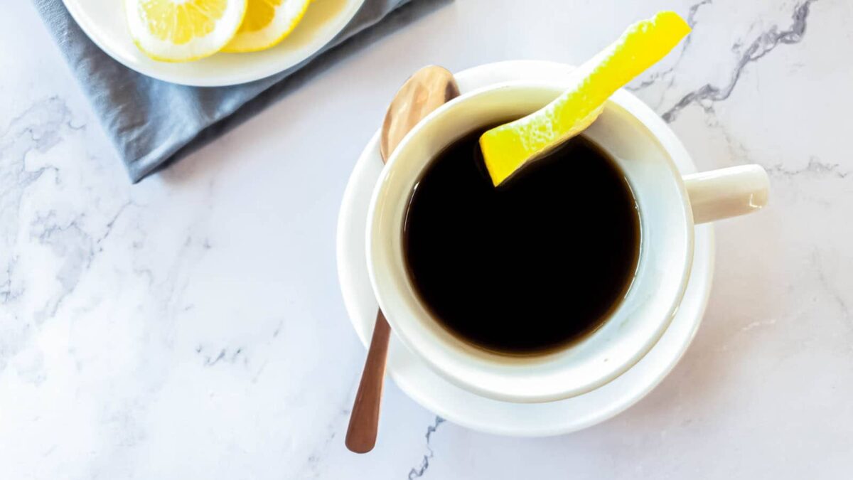 A cup of black coffee with a lemon peel garnish sits on a white saucer with a spoon, next to a plate of lemon slices on a marble countertop—perfect for those who enjoy vibrant sips and vegan drinks made with fresh ingredients.