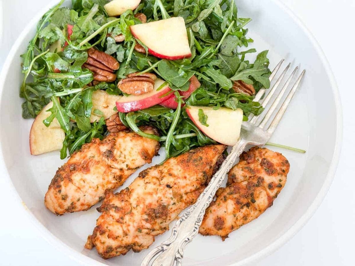 Grilled chicken tenders served with a fresh salad of arugula, apple slices, and pecans, all drizzled with creamy herbs dressing on a white plate with a fork.