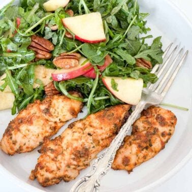 Grilled chicken tenders served with a fresh salad of arugula, apple slices, and pecans, all drizzled with creamy herbs dressing on a white plate with a fork.