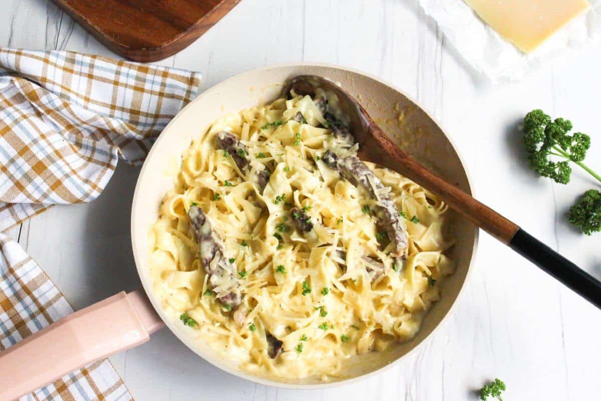 A skillet filled with creamy pasta and mushrooms, garnished with chopped parsley, sits on a white surface next to a wooden spoon, a checkered cloth, and a wedge of cheese—perfect comfort food for fans of classic beef recipes.