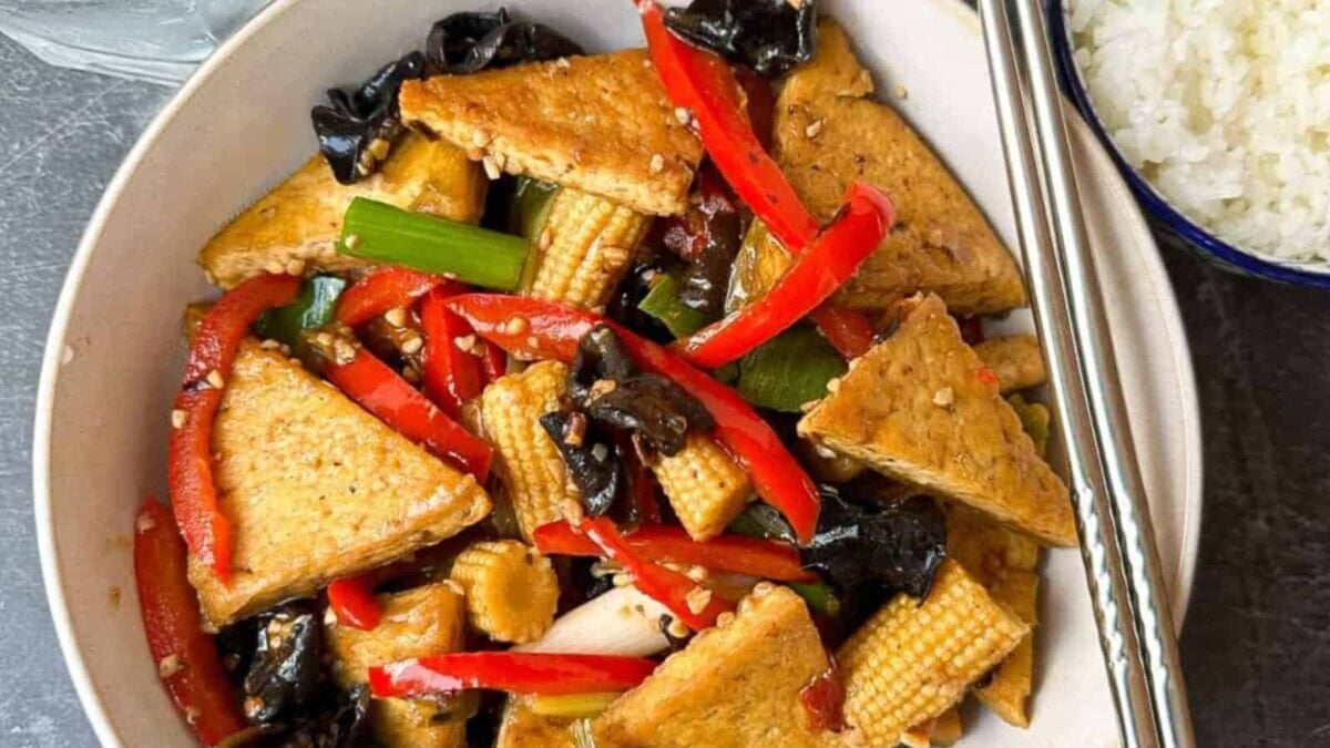 A bowl of stir-fried tofu with baby corn, red bell pepper, black mushrooms, green onions, and savory sauce delivers rich flavor. Served with white rice and metal chopsticks—perfect for fans of delicious tofu recipes.
