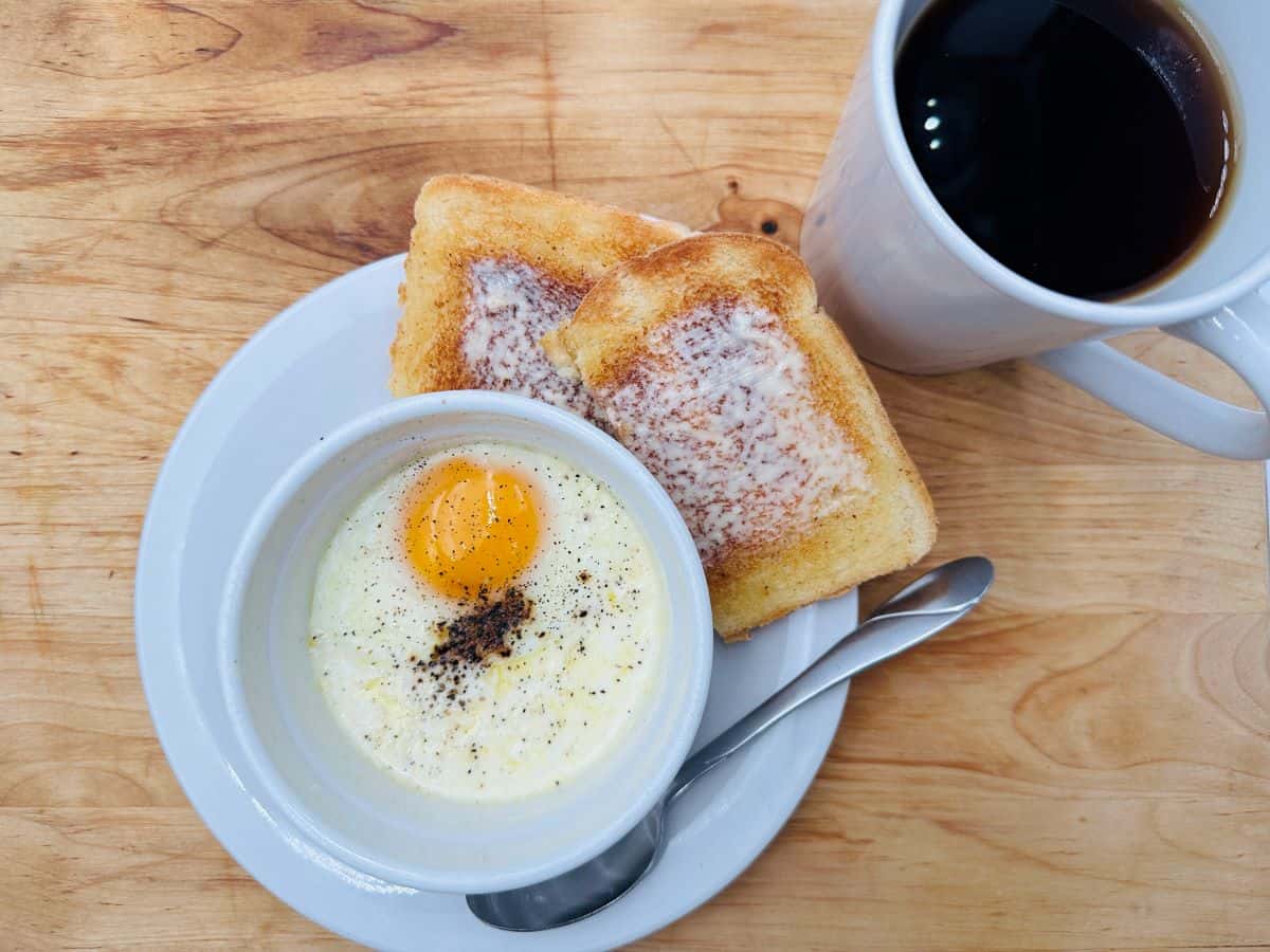 A plate with buttered toast, a baked egg in a ramekin topped with pepper, a spoon, and a cup of black coffee on a wooden table—perfect for those seeking filling morning meals.