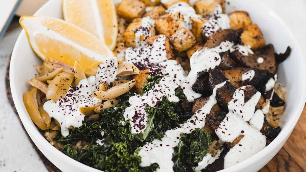 A bowl of rice topped with kale, roasted tofu for silky protein, mushrooms, artichoke hearts, lemon wedges, and a drizzle of white sauce makes for a flavorful twist on classic tofu recipes.
