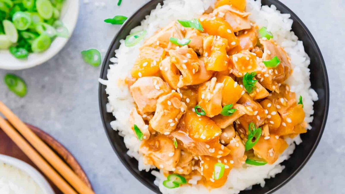 A bowl of white rice topped with diced chicken, pineapple chunks, sesame seeds, and sliced green onions—perfect for fans of flavorful slow cooker recipes. A side dish of sliced green onions and chopsticks are nearby.