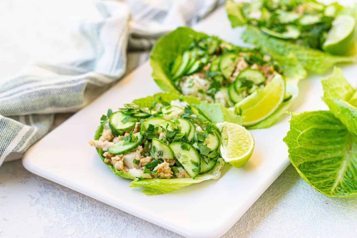 Lettuce leaves topped with diced chicken, sliced cucumber, herbs, and lime wedges create fresh wraps with savory layers on a white rectangular plate.