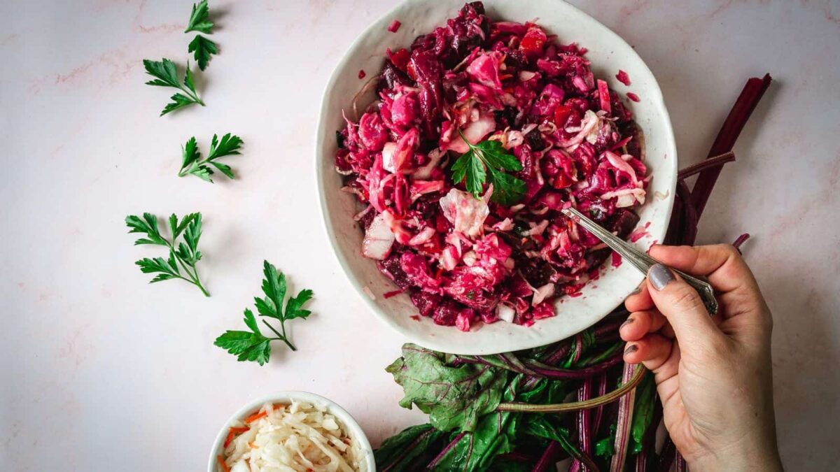 A hand holding a fork over a bowl of chopped beet salad, garnished with parsley, showcases classic flavor in regional cuisine, with fresh greens and a small bowl of shredded cabbage nearby.