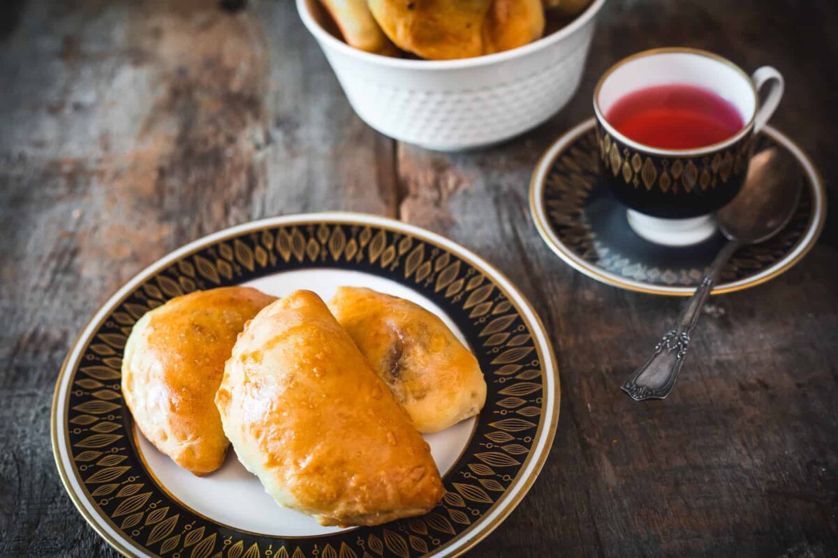 A plate with three baked pastries sits next to a cup of red tea on a saucer, evoking classic flavor found in European dishes, with more pastries in a white bowl in the background on a wooden table.