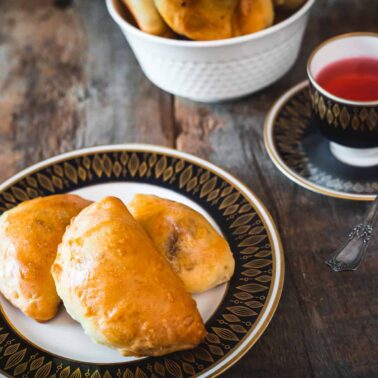 A plate with three baked pastries sits next to a cup of red tea on a saucer, evoking classic flavor found in European dishes, with more pastries in a white bowl in the background on a wooden table.